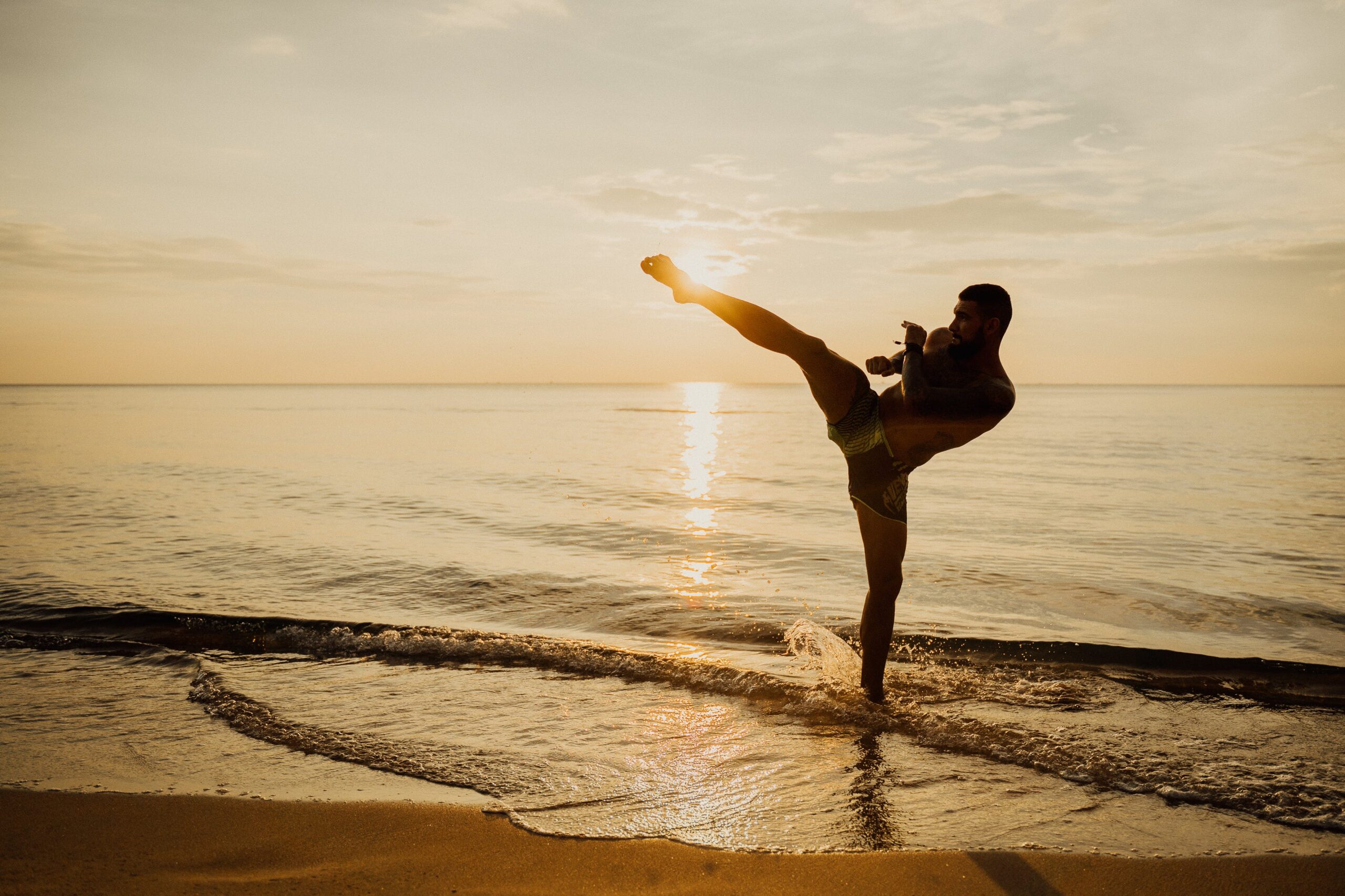 Muay Thai fighter performing a high kick on a tropical beach during a Fight & Travel training camp in Thailand.